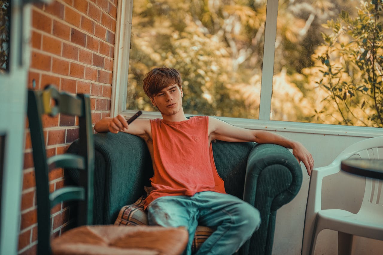 Home A young man in an orange shirt relaxes on a sofa indoors, holding a vape, near a window with a cozy autumn view.