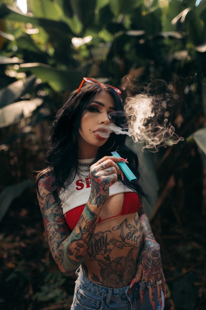 Home Tattooed woman smoking an e-cigarette in a lush outdoor setting with sunglasses and vibrant tattoos.