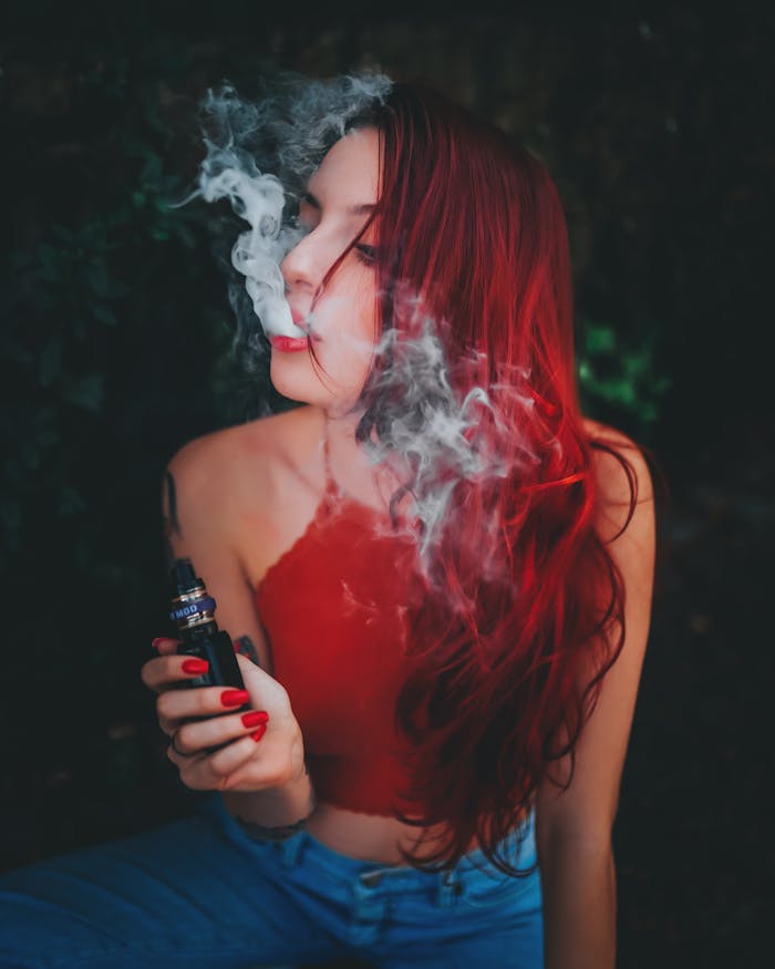 Home Young woman with vibrant red hair uses a vape device, surrounded by smoke in an outdoor setting.